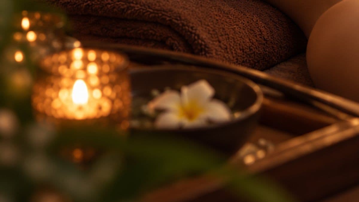 Ánh nến trong cốc thủy tinh cạnh bát nước thả hoa sứ trắng bên giường massage