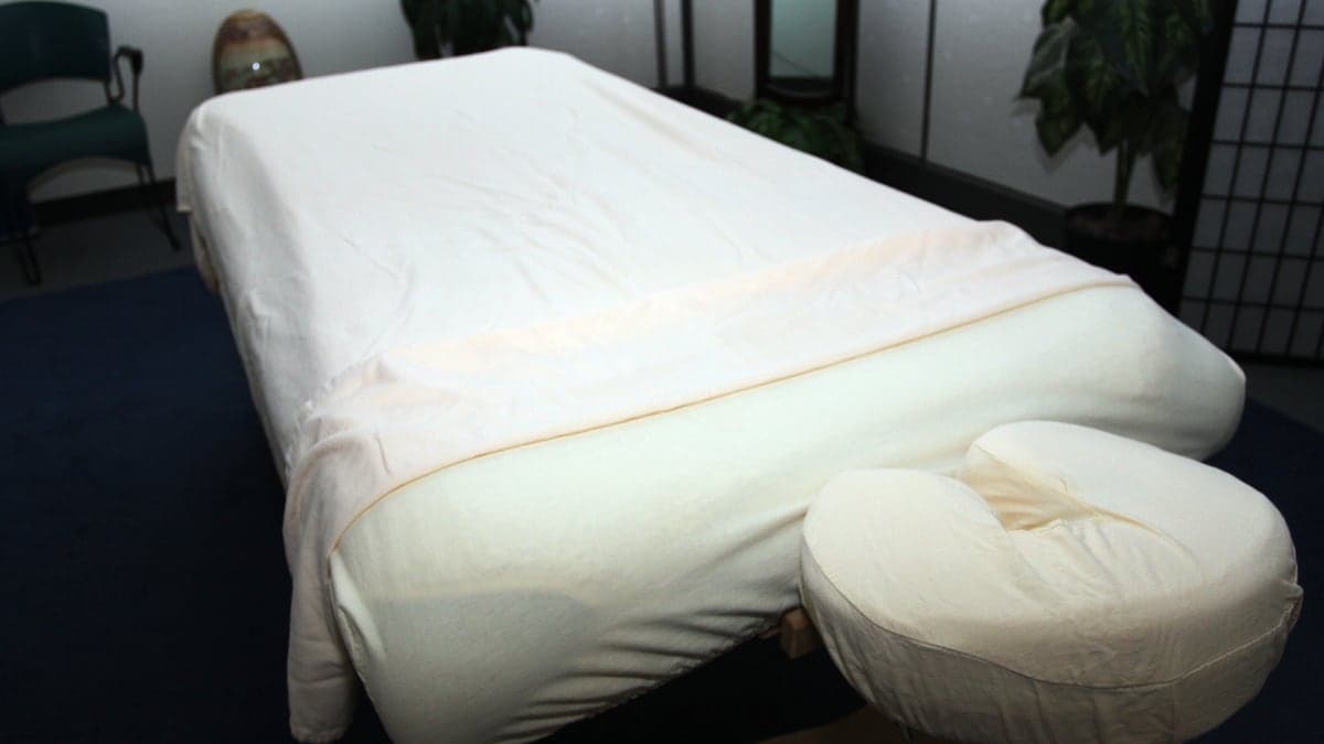 Giường massage trải khăn trắng, có gối tựa mặt, trong một căn phòng có chậu cây