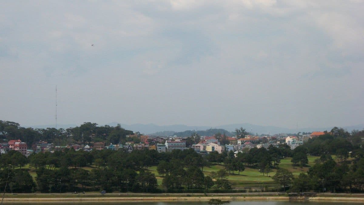 A hazy view across a lake and green lawns to the rooftops and pine-covered hills of Da Lat
